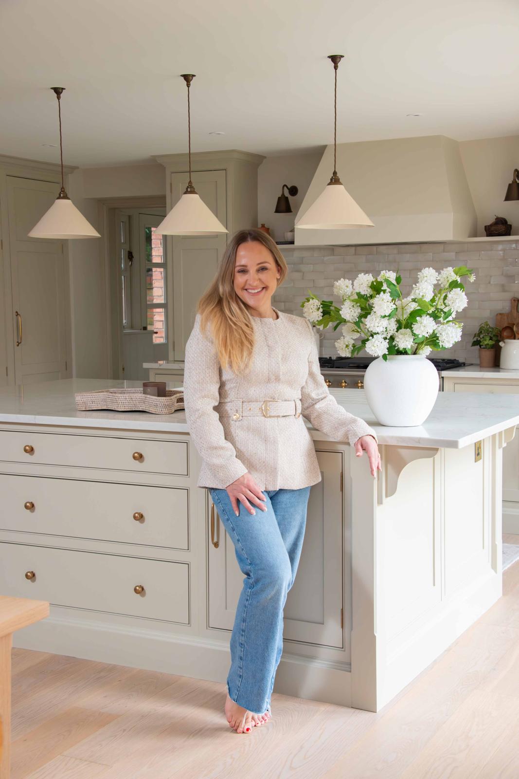Woman standing in a kitchen with light-colored cabinets and pendant lights.